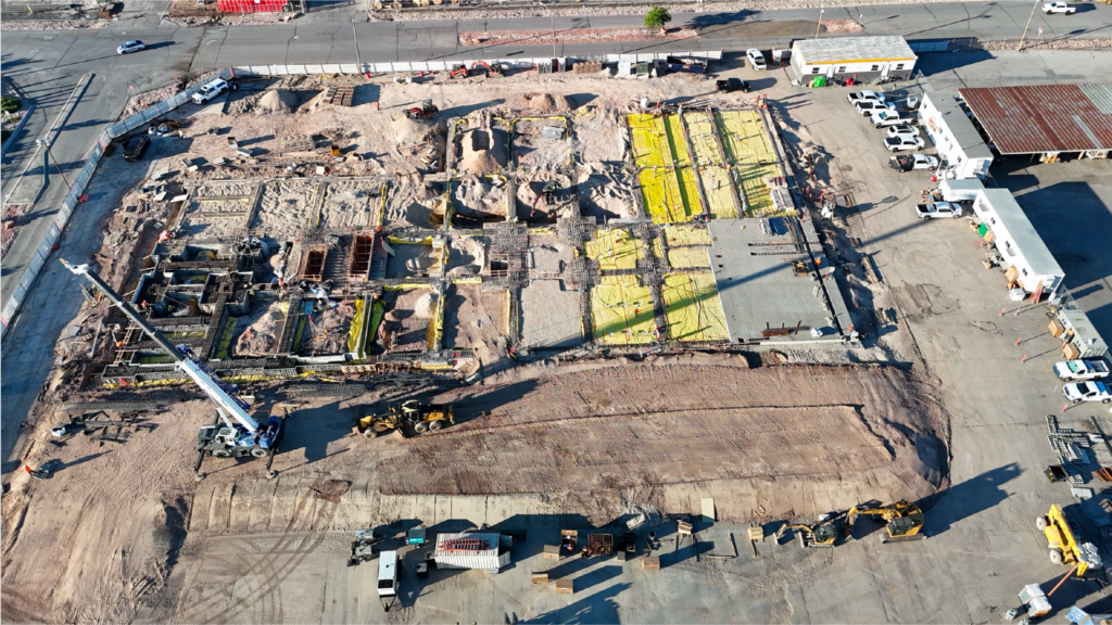Vista aérea de un sitio de construcción con trabajos de cimentación, vehículos de construcción, materiales y estructuras temporales visibles.